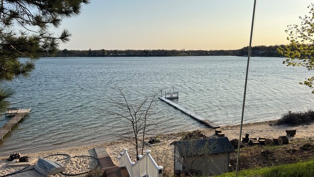 A lakeside view with a dock, a small shed, and a wooden structure on the sand. - Eagles Landing - Lakefront Home on crystal clear Wolf Lake (Muskegon)