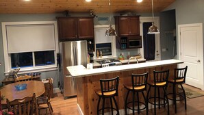 A kitchen with wooden cabinets, a stainless steel refrigerator, a microwave, and a bar area with stools. - Eagles Landing - Lakefront Home on crystal clear Wolf Lake (Muskegon)