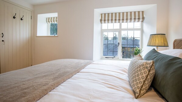 A bedroom with a large bed, a window with a striped valance, a bedside lamp, and a view of the outside. - Oak Cottage - Pet friendly, Swerford (Swerford)