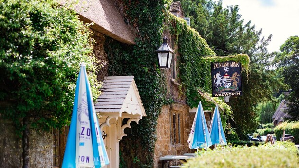 En byggnad med murgrönebeklädda väggar, en skylt med texten ”Falkland Arms WADWORTH” och blåa parasoller.