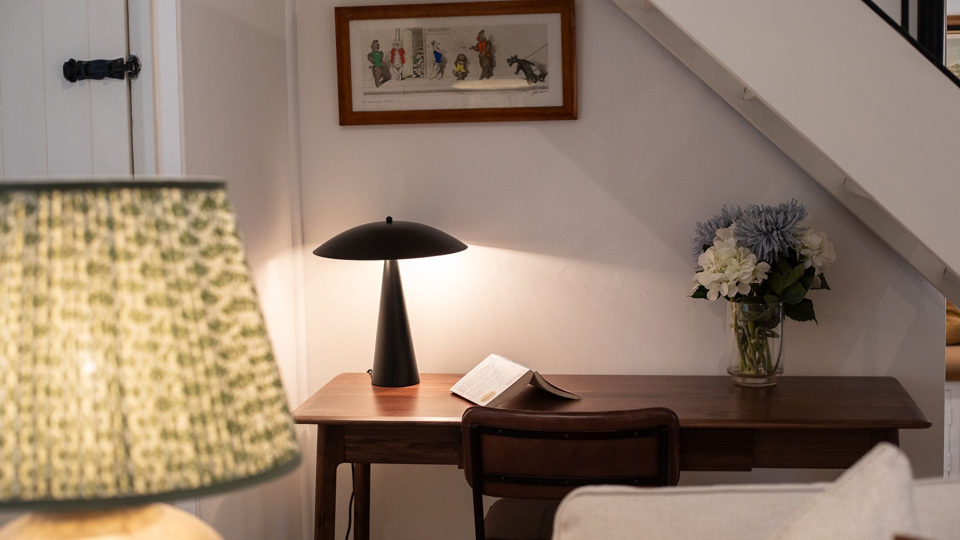 Un bureau en bois, une lampe, une chaise, un vase avec des fleurs et un tableau encadré accroché au mur.