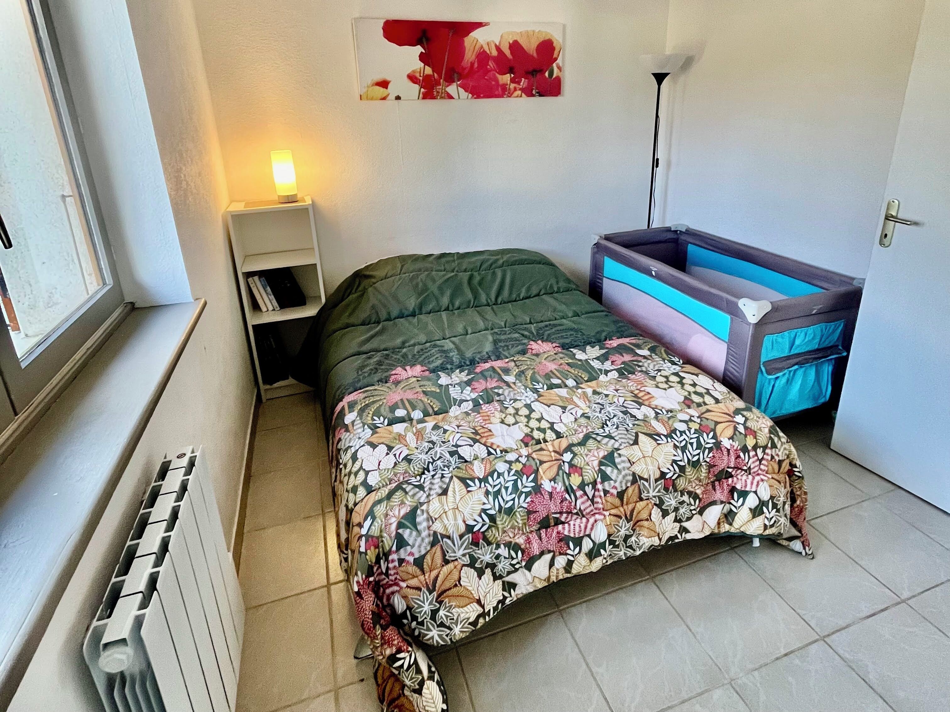 A bedroom with a bed, a nightstand, a radiator, a window, a wall-mounted shelf, and a small storage unit.