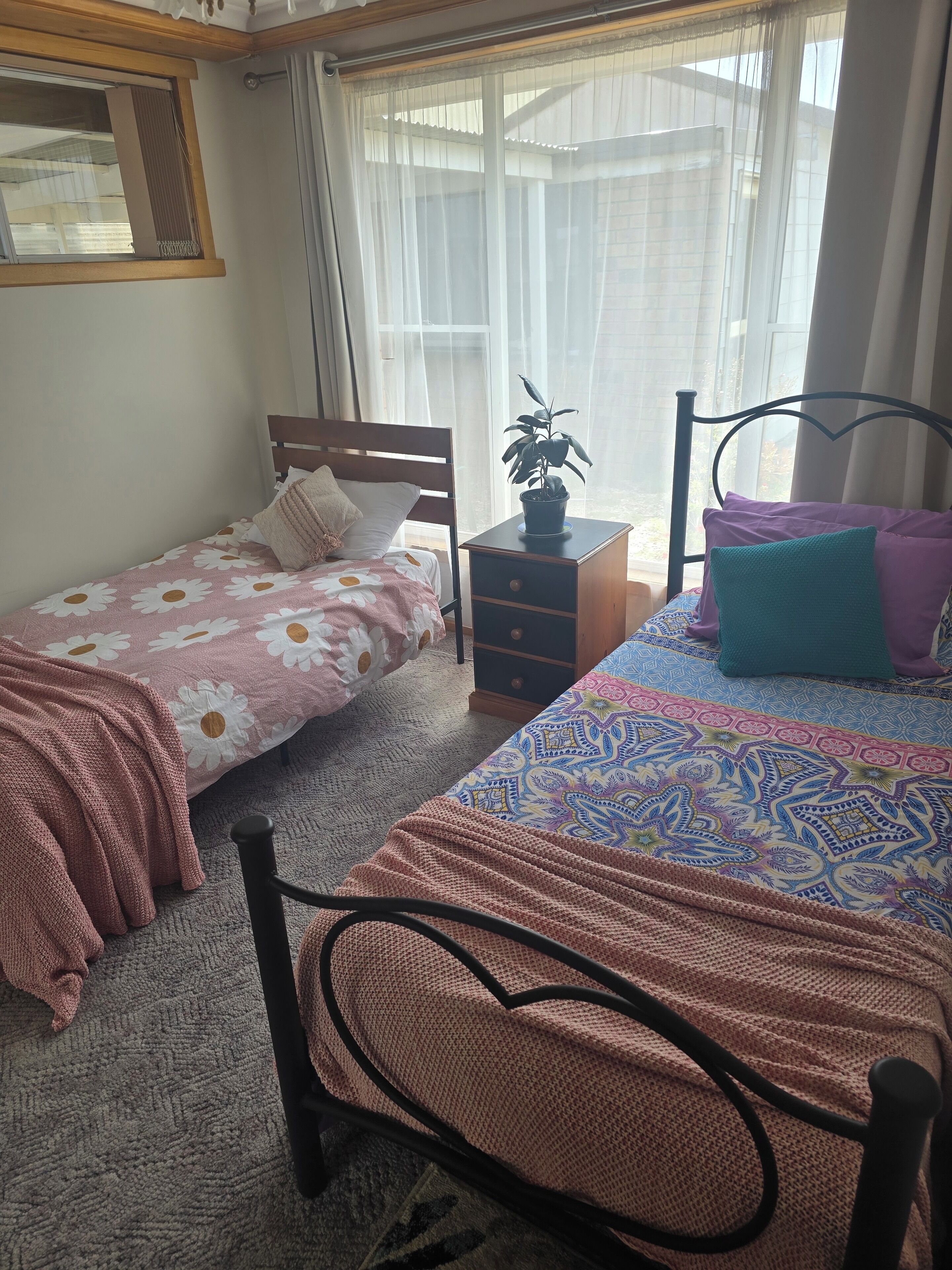 Ein Schlafzimmer mit zwei Einzelbetten, einem Nachttisch, einer Topfpflanze und einem Fenster mit durchsichtigen Vorhängen.
