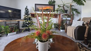 A living room with a round wooden coffee table, a Christmas tree, a large plant, a bookshelf, and a television.