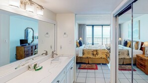 Hotelkamer met een grote spiegel, een badkamer met wastafel en toiletartikelen, een bed met een beddeken met zonmotief, en een balkon met uitzicht.