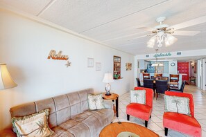 A living room with a brown sofa, red chairs, a ceiling fan, and a kitchen in the background. - Coastal 2BR, Steps to Gulf: Suntide III - Unit 709 (South Padre Island)