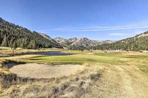 Un parcours de golf avec un étang, entouré de montagnes boisées et offrant une vue sur un ciel dégagé.