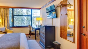 A hotel room with a bed, a desk, a dresser, a television, and a large window with a view of trees. - Everline Resort Studio w/ Luxury Resort Perks (Olympic Valley)