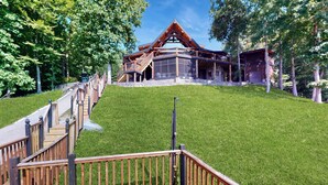 Un chalet moderne doté d’une terrasse en bois menant à une entrée en pierre, le tout entouré d’une végétation luxuriante.