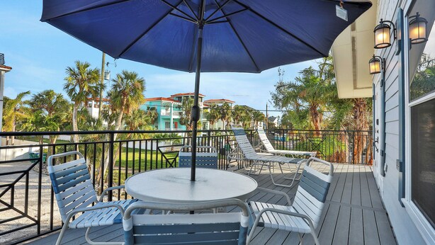 A covered outdoor seating area with a table and chairs, surrounded by a railing and palm trees. - Anna Maria Island Duplex: Pool, Kitchen, Sun Deck (Holmes Beach)