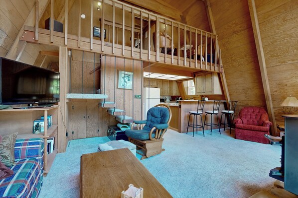 A spacious loft-style room with a high ceiling, wooden beams, a dining area, a kitchen, and a cozy seating area with a sofa and a chair. - Sierra Nevada Hideaway: Cabin w/ Deck, Mtn Views (Graeagle)