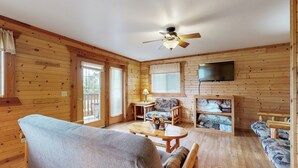 A wooden cabin interior with a sofa, a small table, a chair, a television mounted on the wall, and a ceiling fan. - Adventure Basecamp! Three Bears Resort Retreat (Warrens)