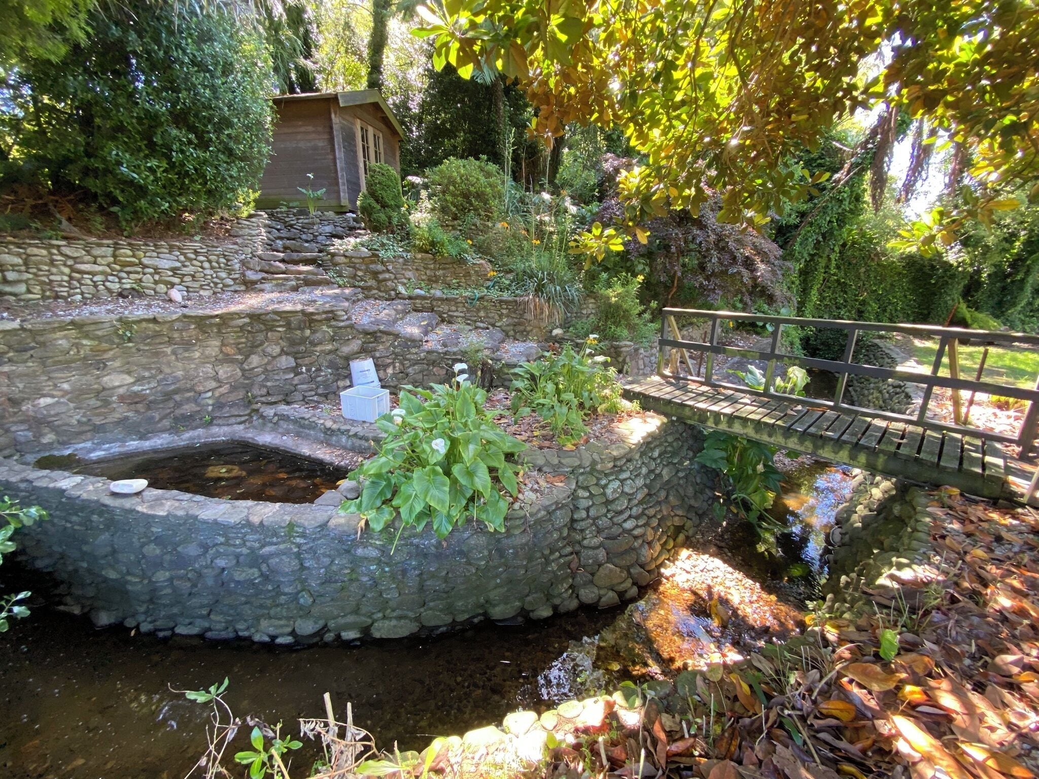 Un bassin en pierre, un pont en bois et une petite construction en bois dans un jardin.