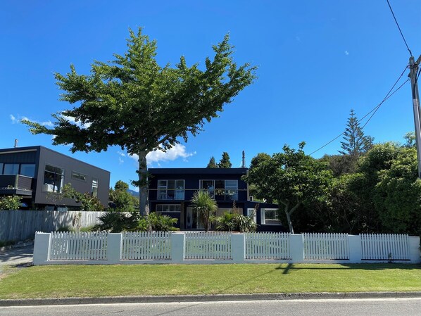 Una casa moderna de dos pisos con cerca blanca de piquete, un árbol grande al frente y un cielo azul despejado.
