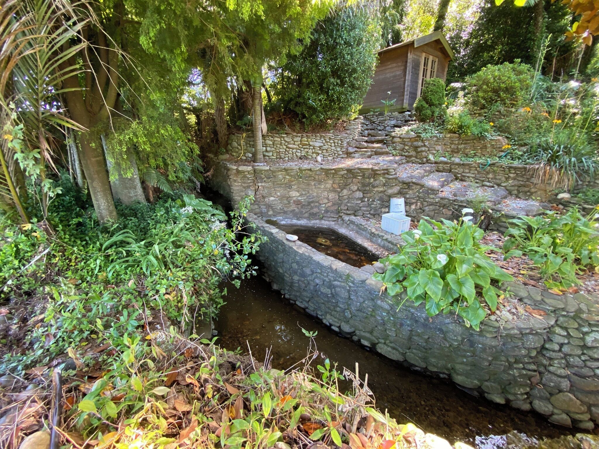 A stone water feature with a small pond, surrounded by lush greenery and a wooden structure.