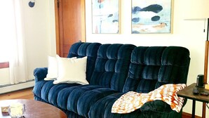A dark blue sofa with a white pillow and a patterned throw blanket, a small wooden table with a bowl and remote control, and abstract art on the wall. - Quiet Downtown Apt w/ Fireplace (Waterville)