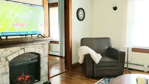 A living room with a stone fireplace, a flat-screen TV mounted above it, a dark leather armchair, a clock on the wall, and a window with blinds. - Quiet Downtown Apt w/ Fireplace (Waterville)