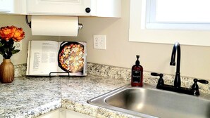 A kitchen with a granite countertop, a stainless steel sink, a red soap dispenser, and a cookbook open to a recipe page. - Quiet Downtown Apt w/ Fireplace (Waterville)