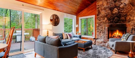 Una sala de estar acogedora con chimenea de piedra, un sofá grande y un comedor con vistas al exterior.