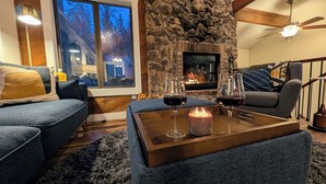 A cozy living room with a stone fireplace, two sofas, a coffee table with glasses and a candle, and a large window overlooking a snowy landscape.