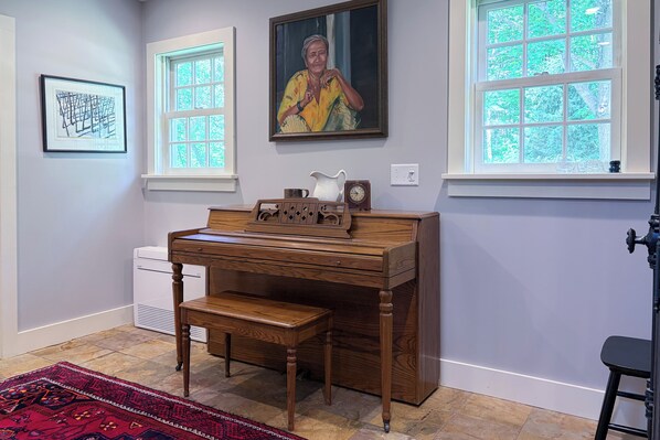 Quarto com escrivaninha de madeira, um banco, um relógio, quadro emoldurado e janela com vista para árvores.