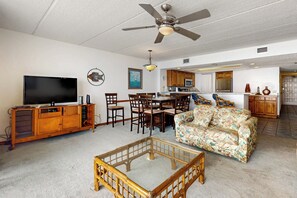 A living room with a TV, wooden furniture, a ceiling fan, and a kitchen area in the background. - Gulf Views, Resort Perks: Suntide III - Unit 703 (South Padre Island)
