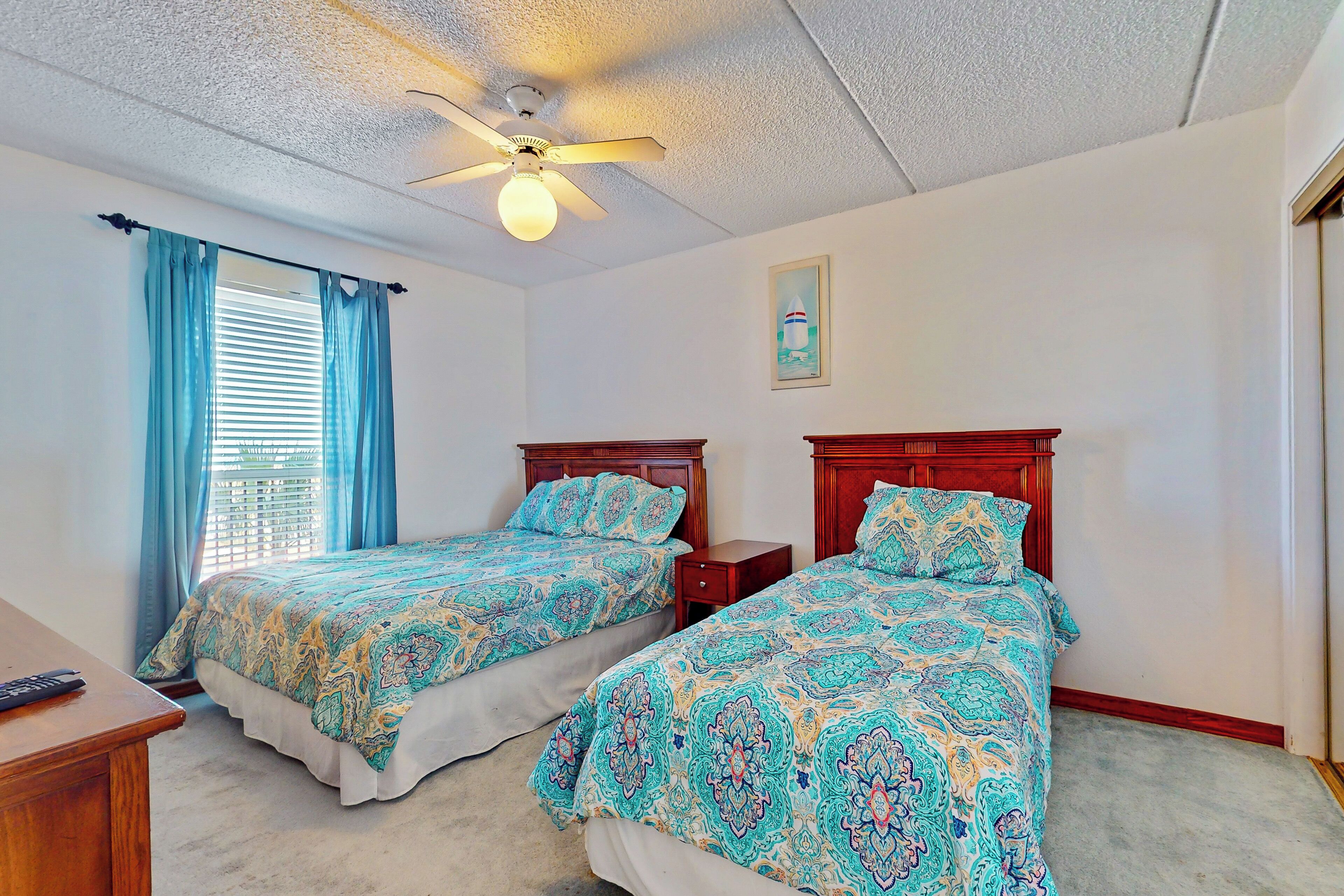A bedroom with two beds, a ceiling fan, a window with curtains, and a small bedside table.
