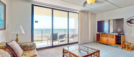 A living room with a sofa, a glass coffee table, a TV on a wooden stand, and a balcony with chairs.