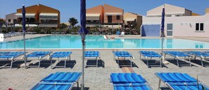 A swimming pool with blue lounge chairs and umbrellas, surrounded by buildings with balconies and striped awnings. - Pine Beach House - Tourist Village with Pool and Animation (Sicilia)
