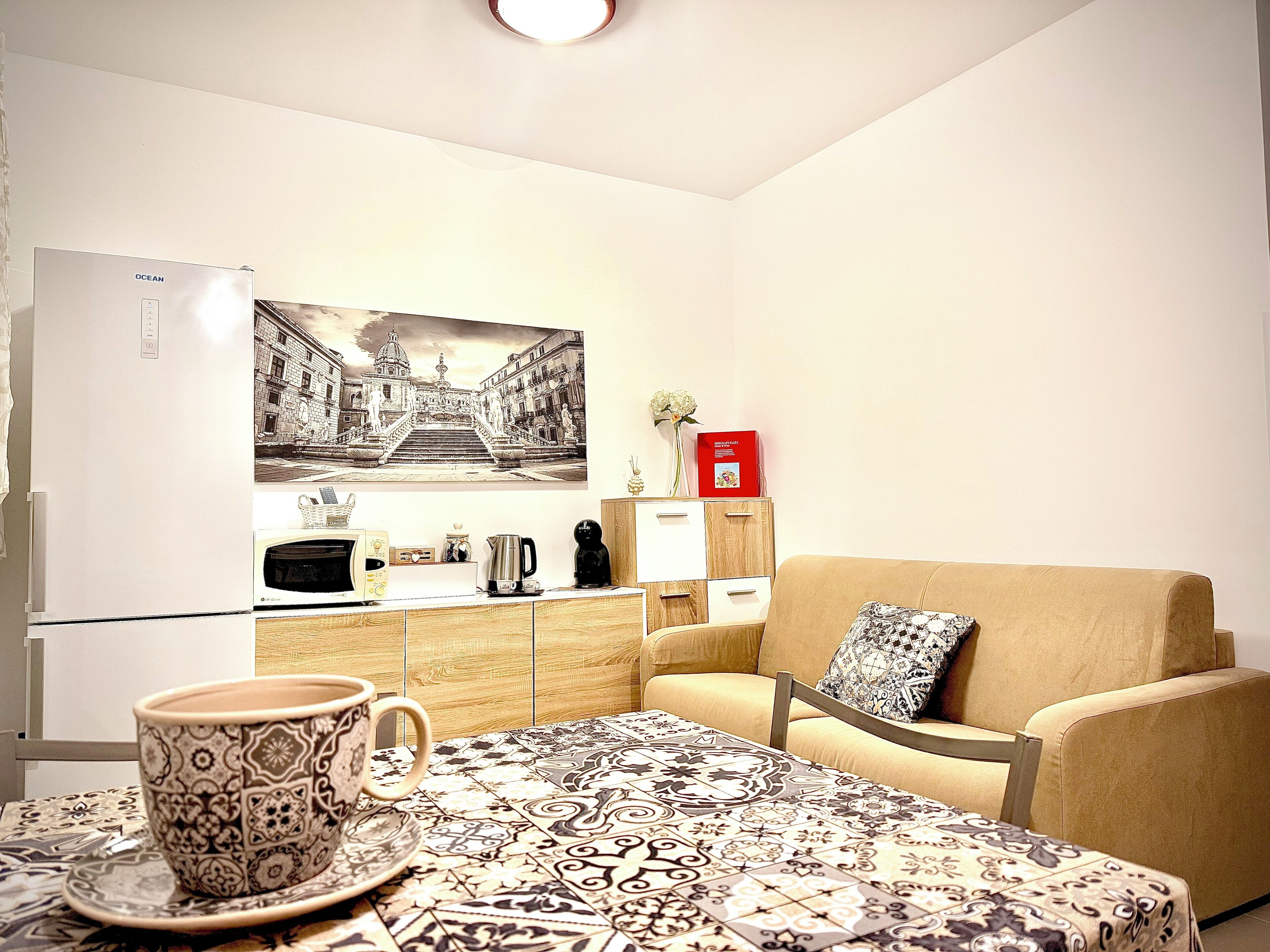 A compact living space with a kitchenette, a dining area with a patterned tablecloth, a sofa with a patterned cushion, and a wall-mounted picture.