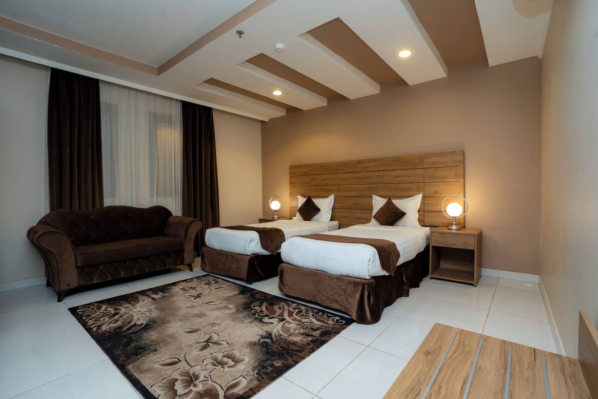 A hotel room with a sofa, two beds, a nightstand with a lamp, and a floral rug.