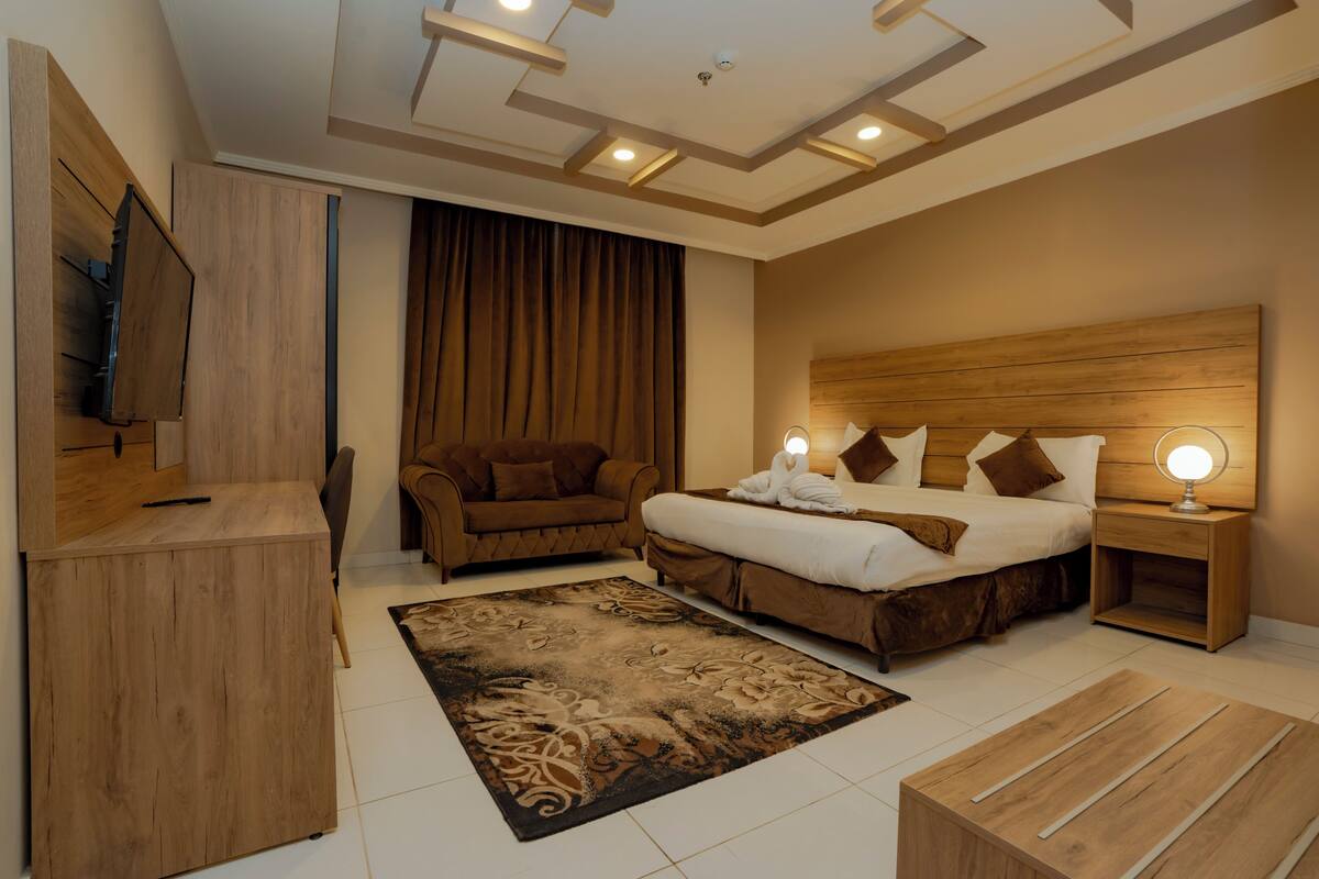 A modern hotel room with a wooden headboard bed, a sofa, a desk, and a TV.