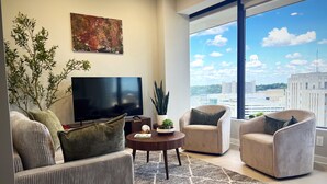 Ein modernes Wohnzimmer mit einem großen Flachbildfernseher, bequemen Sitzgelegenheiten, einem Couchtisch und Blick auf die Stadt durch große Fenster.