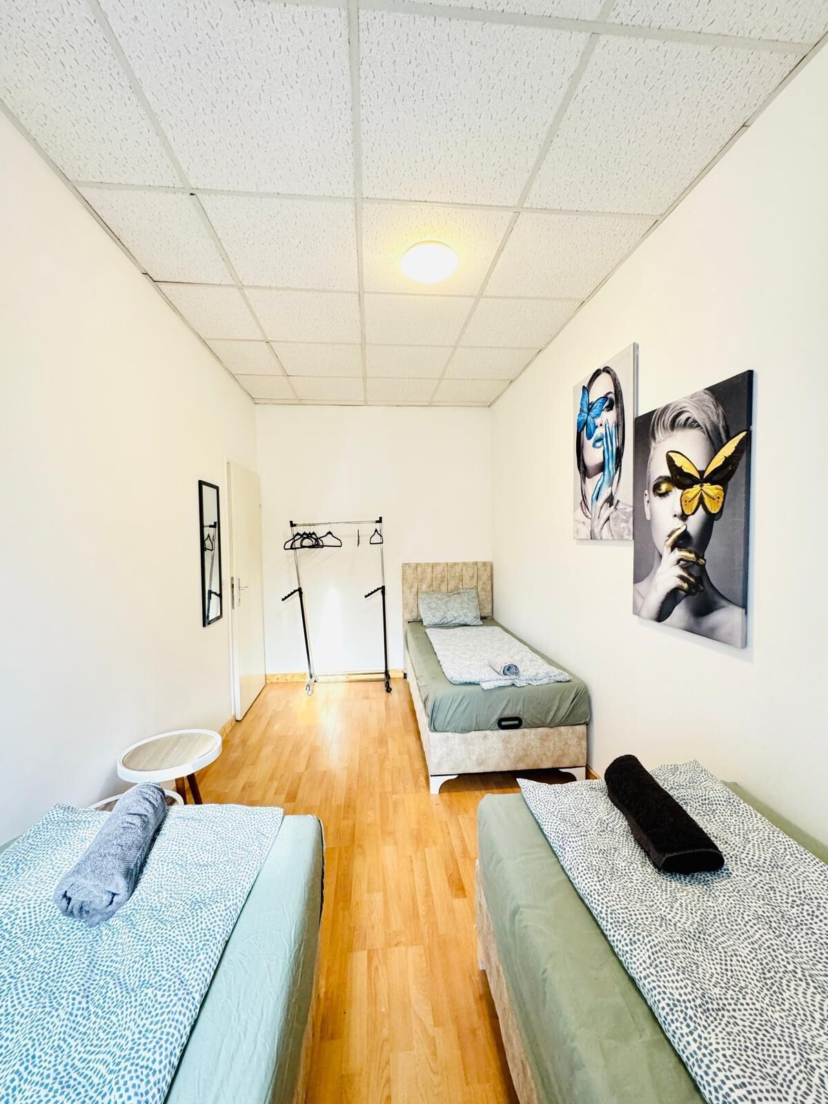 A small, well-lit room with two beds, a small table, and artwork on the walls.