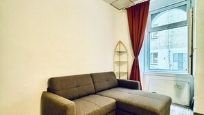 Un salon moderne avec un canapé d’angle, un plancher en bois, une grande fenêtre avec des rideaux et un radiateur.