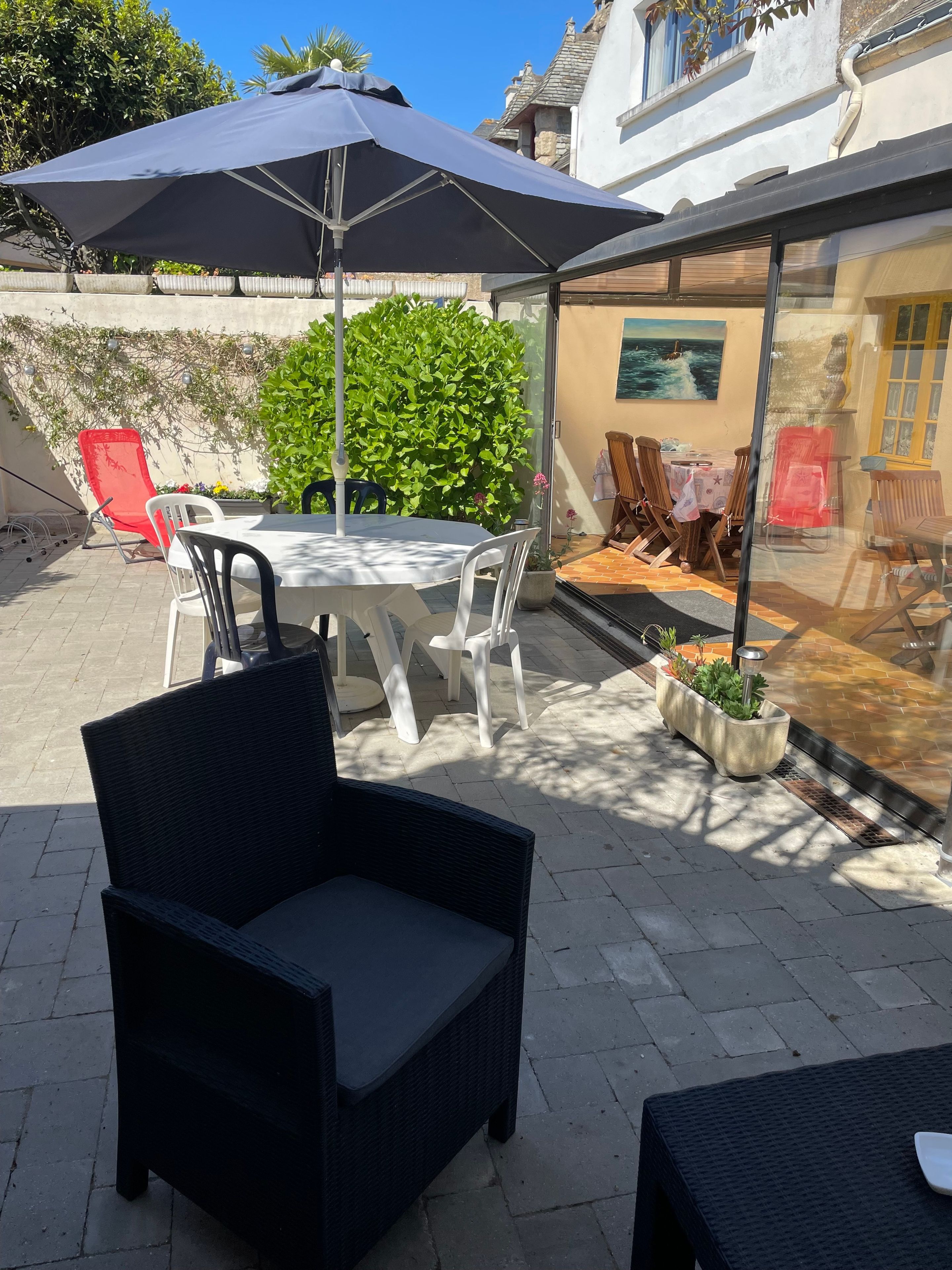 Un patio al aire libre con una mesa, sillas, un paraguas grande y un cerramiento de vidrio que da a un espacio interior.