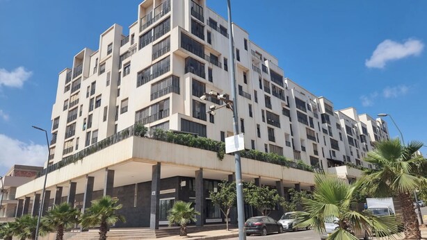 Un immeuble résidentiel à plusieurs étages, doté de balcons, d’une entrée couverte et de palmiers.