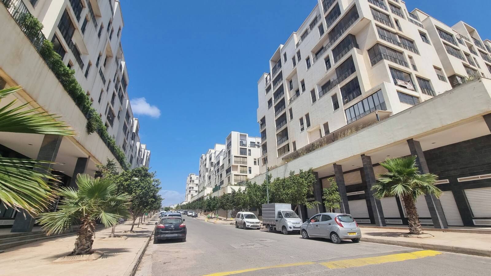 A street lined with multi-story residential buildings, parked cars, and palm trees.