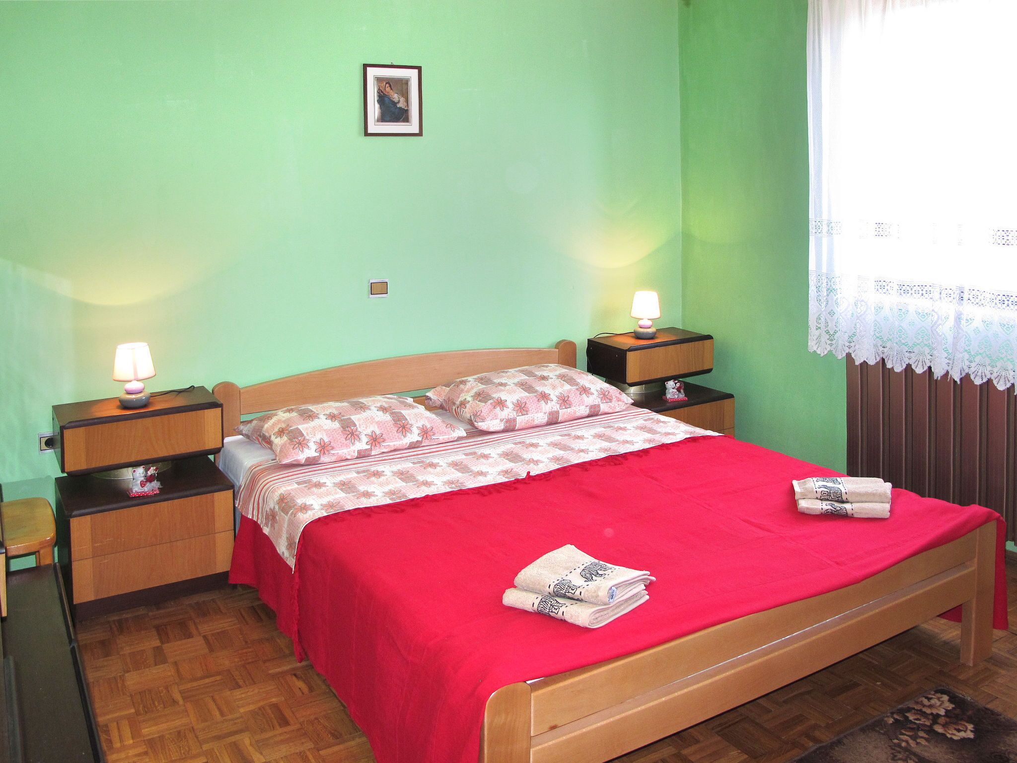 A neatly made bed with a red bedspread and matching pillows, two bedside lamps, a small framed picture on the wall, and a window with a lace curtain.