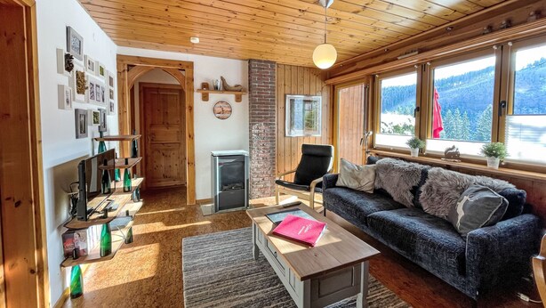 Ein gemütliches Wohnzimmer mit Holzbalkendecke, Kamin, einer Couch, einem Couchtisch und einem Fenster mit Blick auf eine verschneite Landschaft.