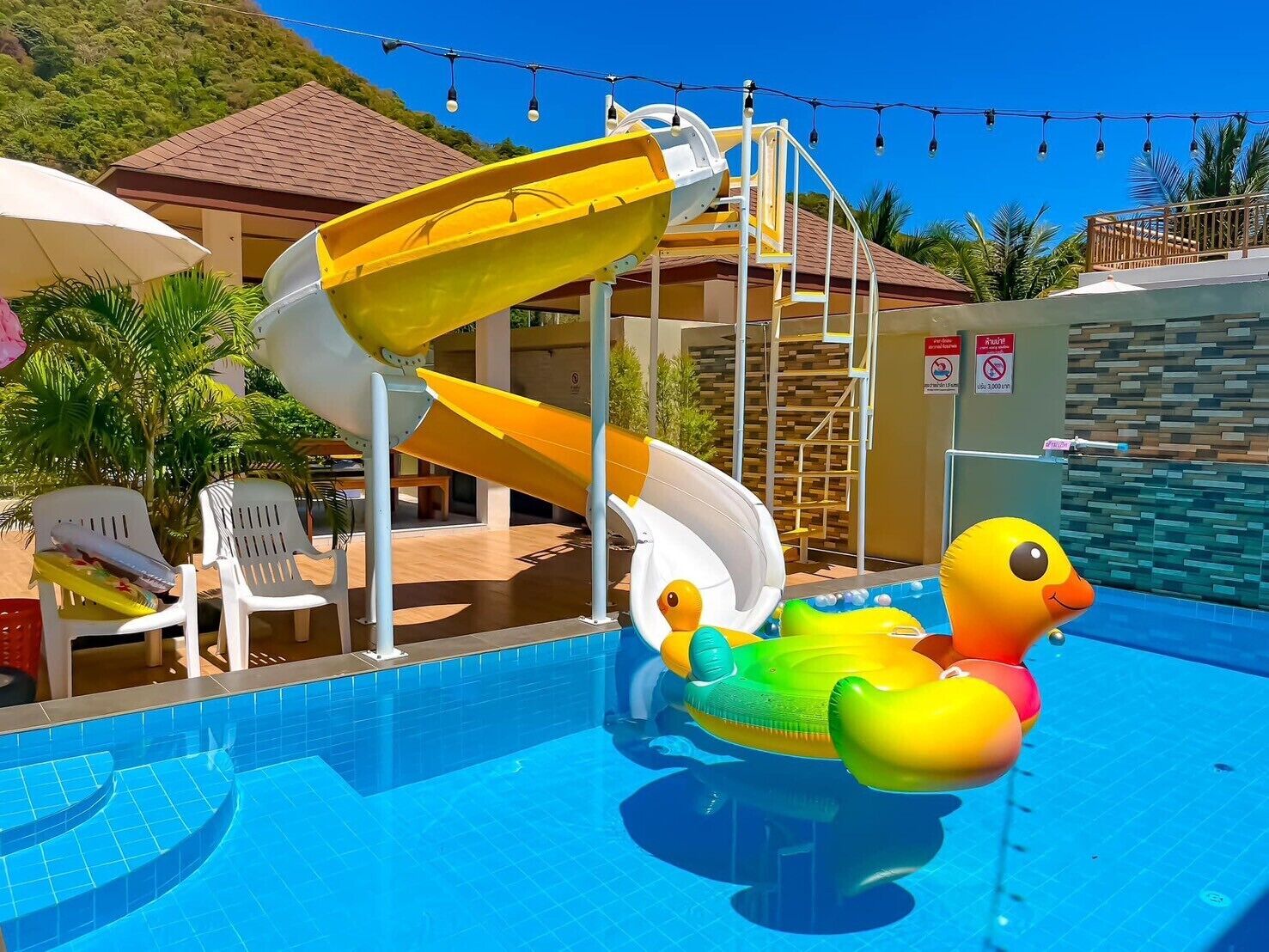 A swimming pool with a yellow and white water slide, inflatable duck float, and a poolside area with chairs and umbrellas.