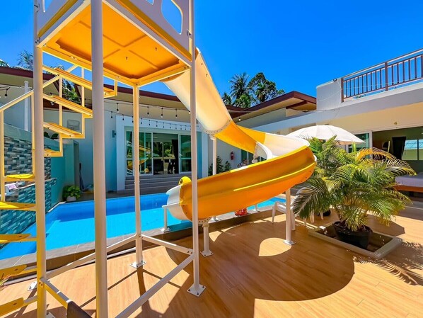 A pool area with a yellow slide, a pool, and a building with a balcony. - Le poulet poolvilla khanom (Khanom)