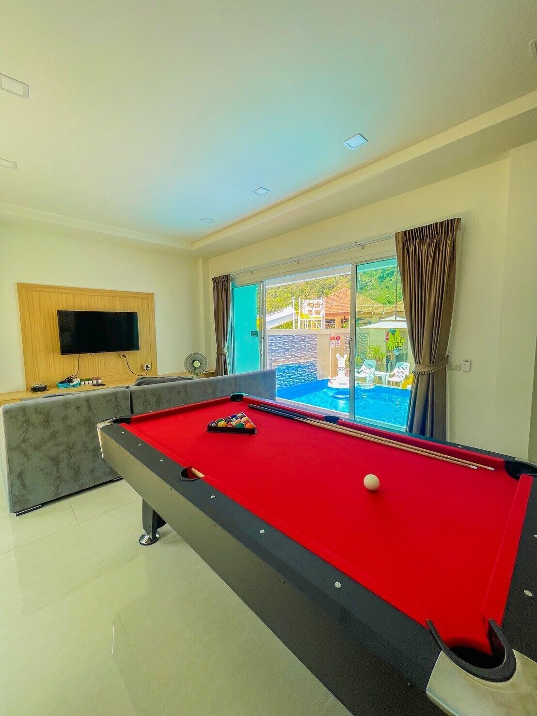 A pool table with a red felt surface, a sofa, a television mounted on the wall, and a view of an outdoor swimming pool through a glass door.