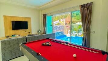 A pool table with a red felt surface, a sofa, a television mounted on the wall, and a view of an outdoor swimming pool through a glass door.