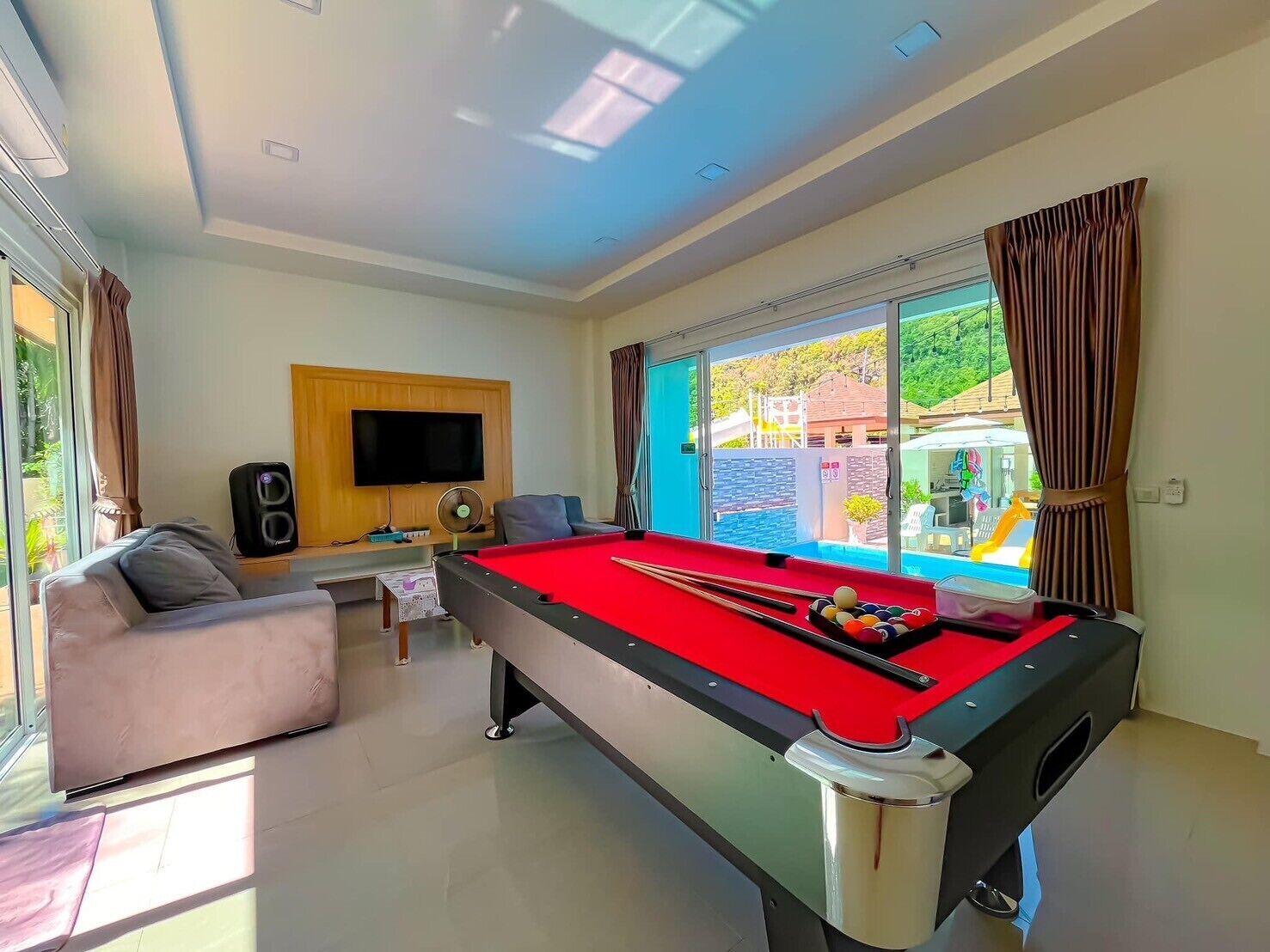 A pool table in a room with a TV, a sofa, and a view of a pool outside.