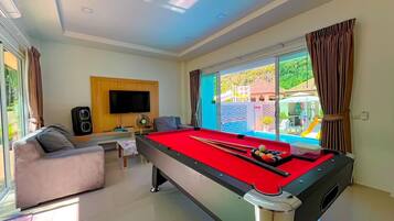 A pool table in a room with a TV, a sofa, and a view of a pool outside.