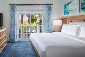 A hotel room with a bed, a TV, a nightstand, a lamp, and a view of palm trees through a sliding glass door.