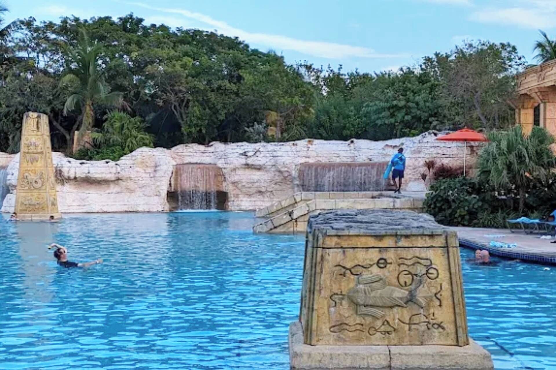 En swimmingpool med en stenskulptur i förgrunden, ett vattenfall och en person som simmar.