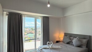 A spacious bedroom with a large bed, a view of the city through floor-to-ceiling windows, and a modern light fixture.