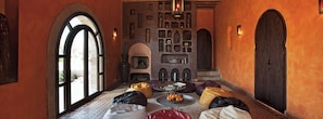 A cozy hallway with a tiled floor, a seating area with cushions, a wall-mounted shelf with decorative items, and a large wooden door. - Villa Gonatouki (ESSAOUIRA)